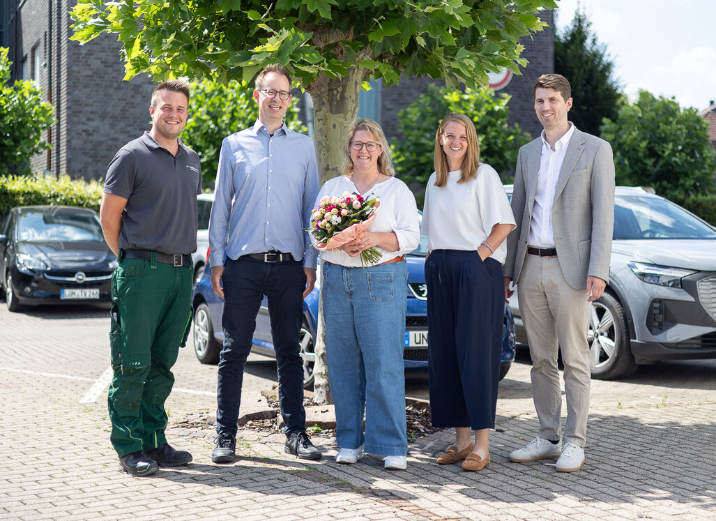 Fünf Mitarbeitende der WBG Lünen stehen unter einem Baum auf einem Parkplatz. Im Mittelpunkt hält eine neue Mitarbeiterin einen Blumenstrauß zur Begrüßung.