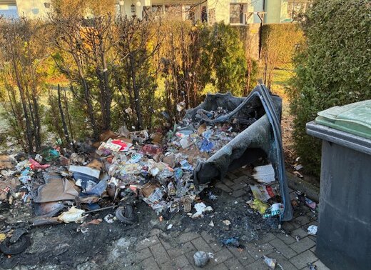 Stark verschmutzter Müllplatz nach einem Mülltonnenbrand: verbrannte Abfälle, geschmolzene Kunststoffreste und umgekippte Mülltonnen liegen vor einer Hecke.