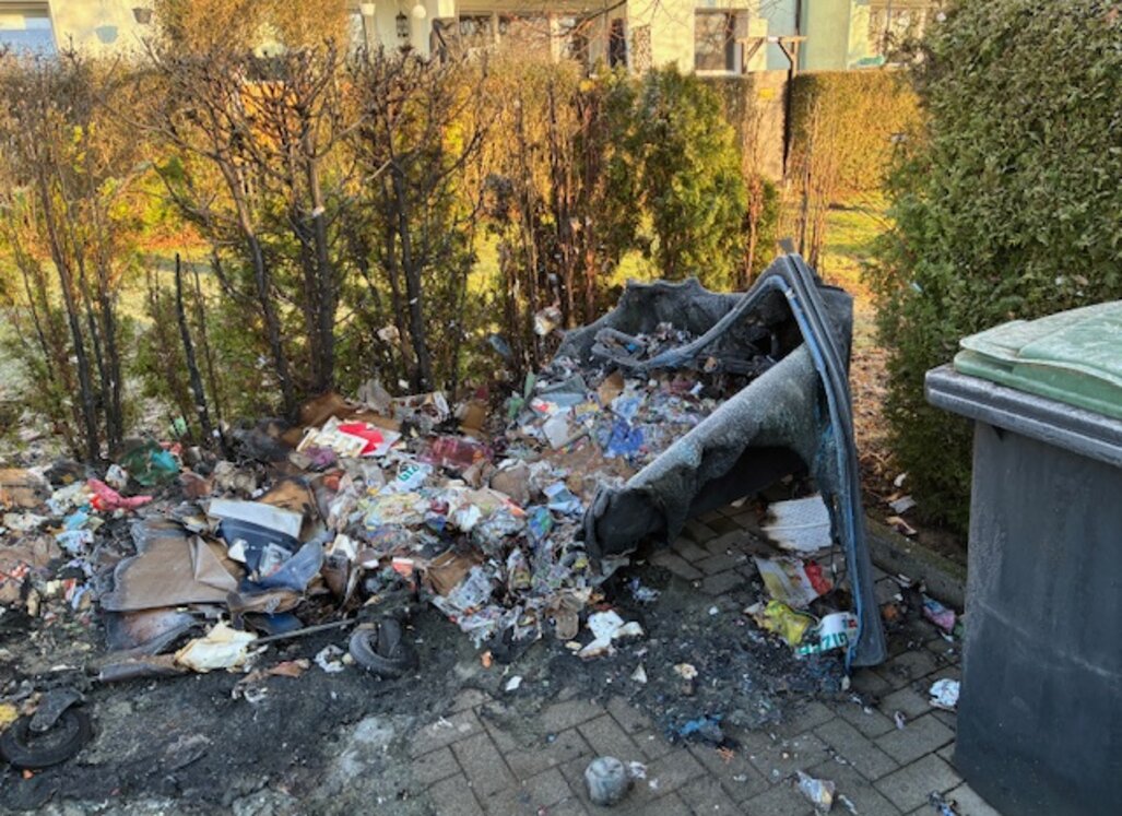 Stark verschmutzter Müllplatz nach einem Mülltonnenbrand: verbrannte Abfälle, geschmolzene Kunststoffreste und umgekippte Mülltonnen liegen vor einer Hecke.