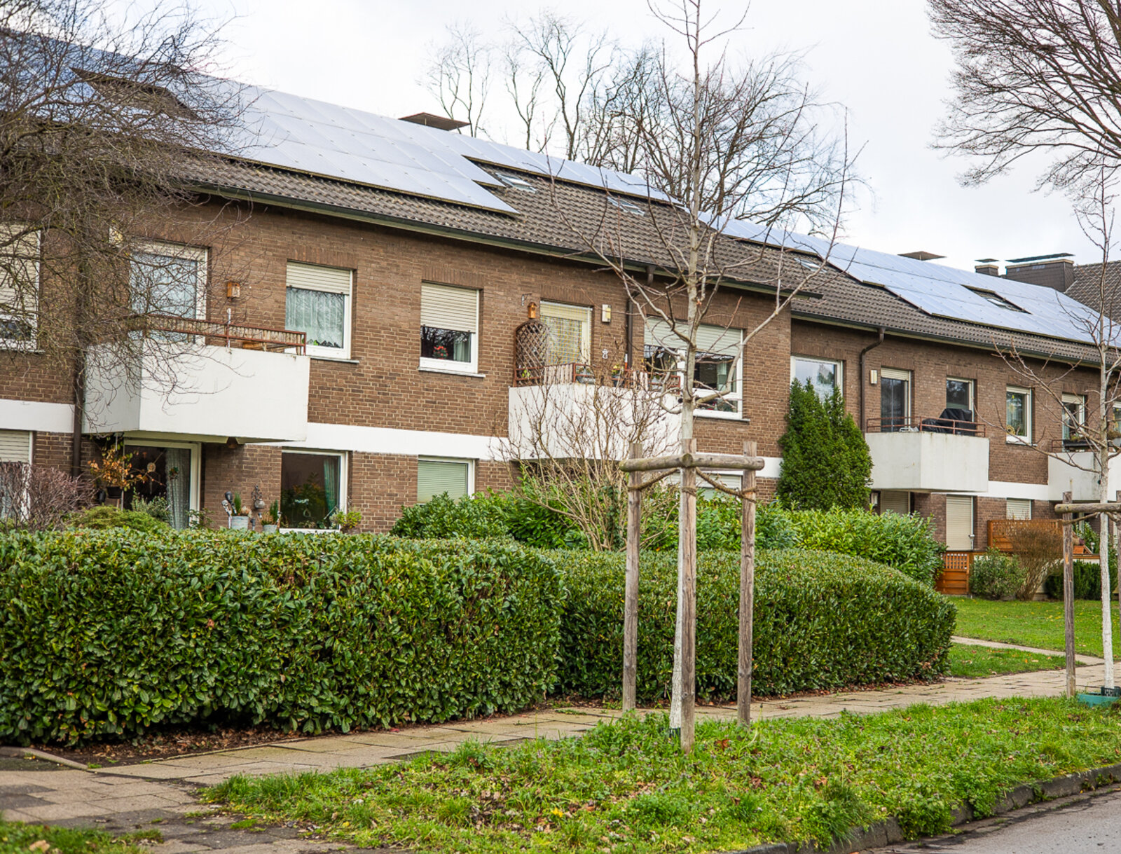 Mehrfamilienhaus mit Klinkerfassade, Balkonen und Solaranlage auf dem Dach, umgeben von Hecken, Bäumen und Grünflächen.