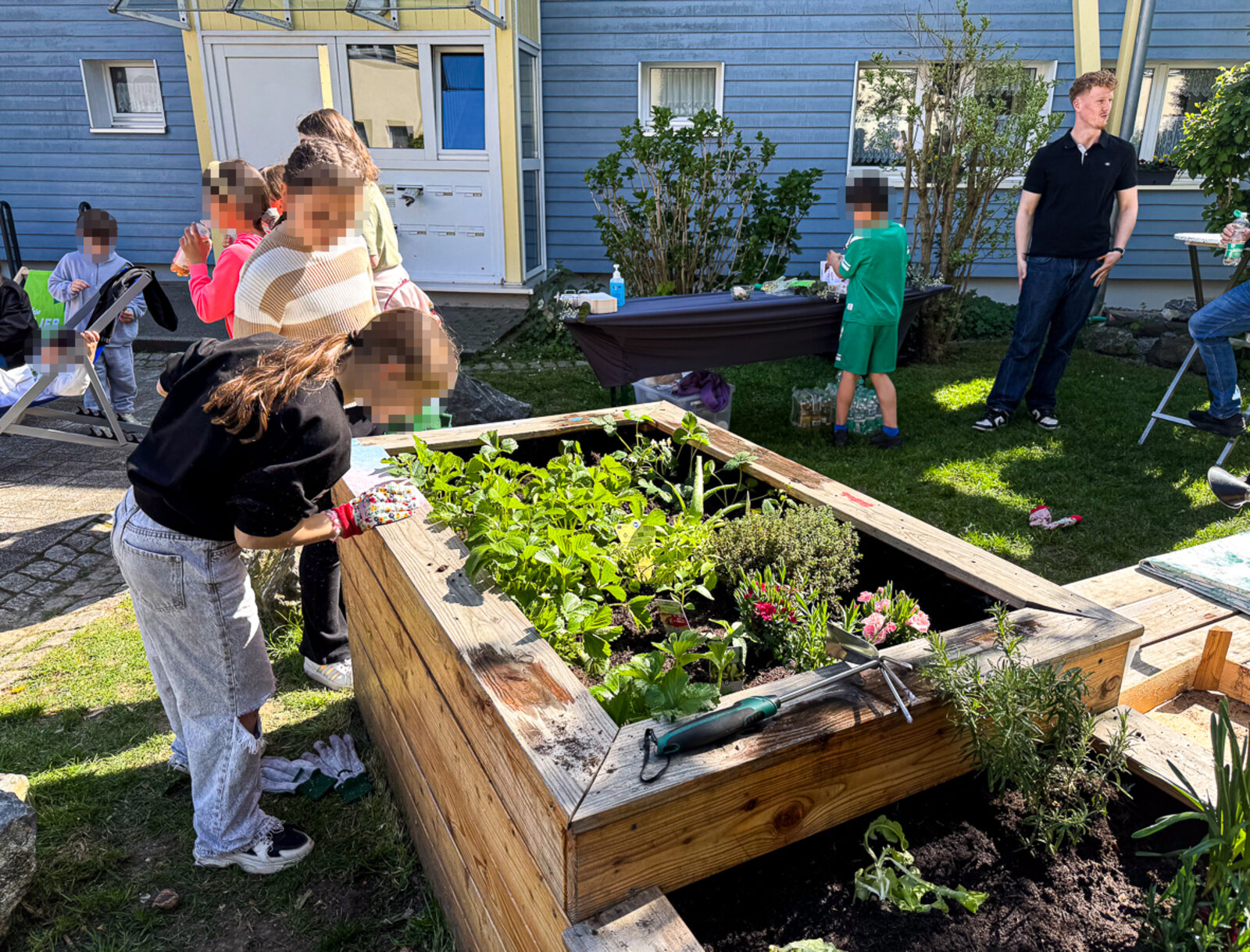 Kinder bepflanzen gemeinsam ein großes Hochbeet mit Kräutern und Blumen, Erwachsene stehen im Hintergrund.
