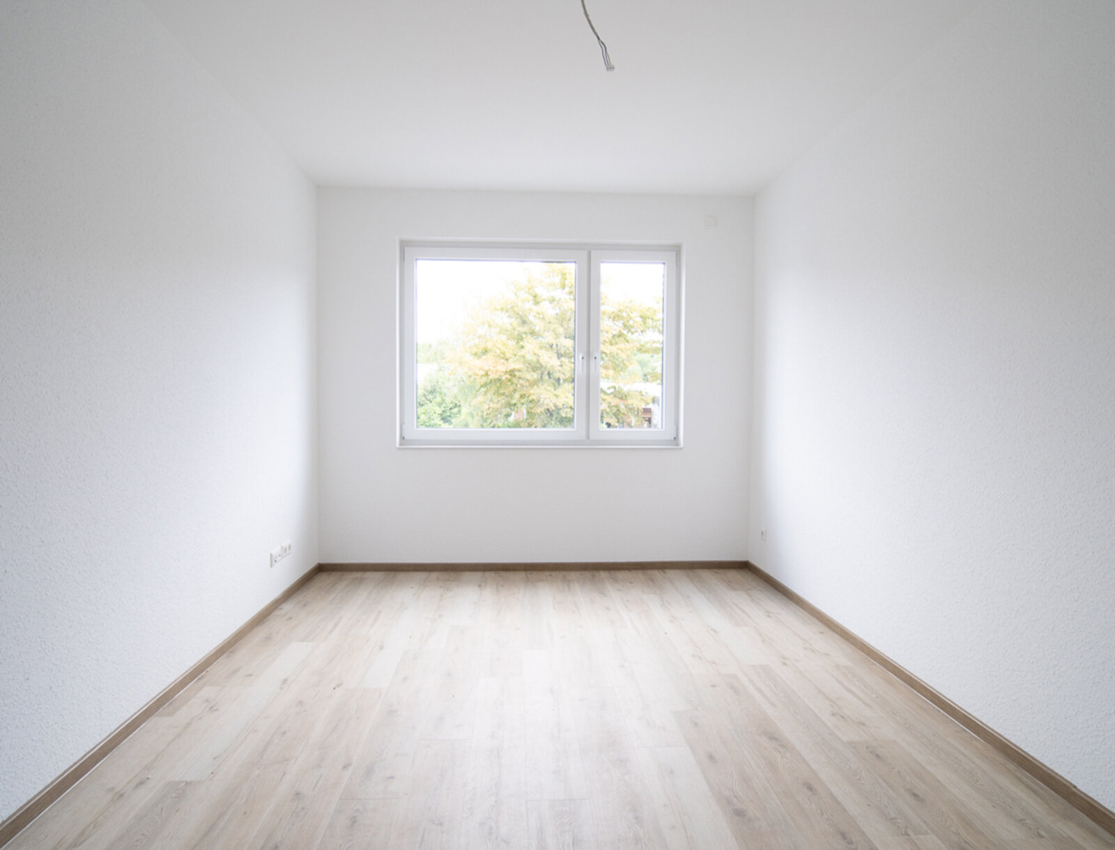 Blick in einen hellen Schlafraum mit gegenüberliegendem Fenster. Ausstattung mit hellem Laminat und weißer Raufasertapete.