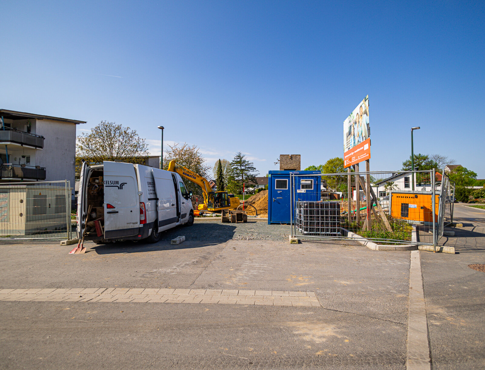 Zufahrt zu der Baustelle mit Bauzaun, Transporter und Baucontainern. Im Hintergrund ein Bagger und ein Bauschild für das Neubauprojekt.