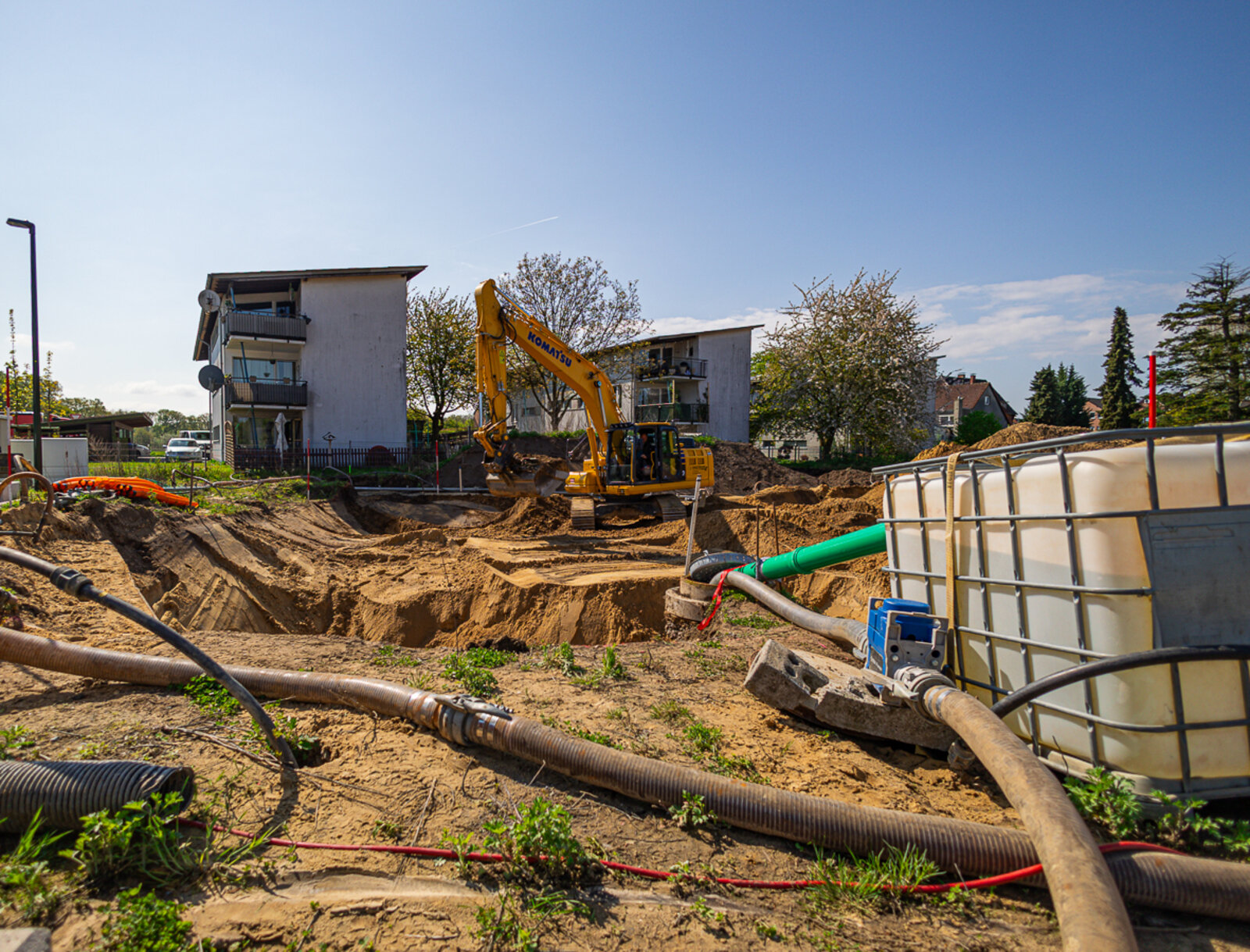 Baustelle mit Baugrube und Bagger im Einsatz. Schläuche und Wasserbehälter liegen im Vordergrund, Wohnhäuser im Hintergrund.