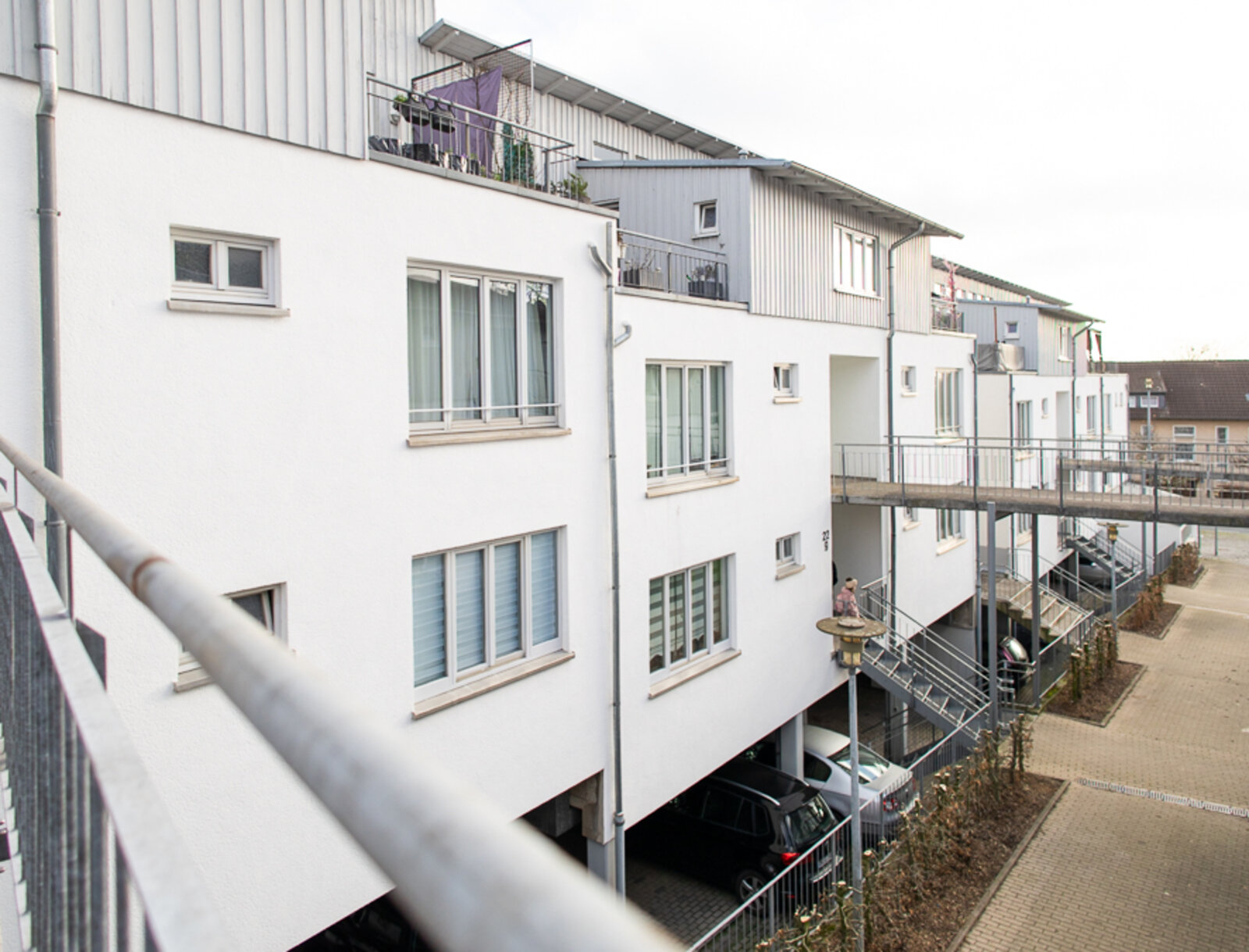 Blick entlang der Wohnanlage mit hellen Fassaden, Balkonen und Außentreppen. Unter den Gebäuden sind Stellplätze zu sehen, daneben gepflasterte Wege und begrünte Bereiche zwischen den Häusern.