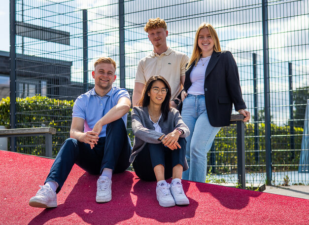 Gruppenfoto: Auszubildende sitzen und stehen gemeinsam vor einem Fußballplatz. Alle Personen lächeln in die Kamera.