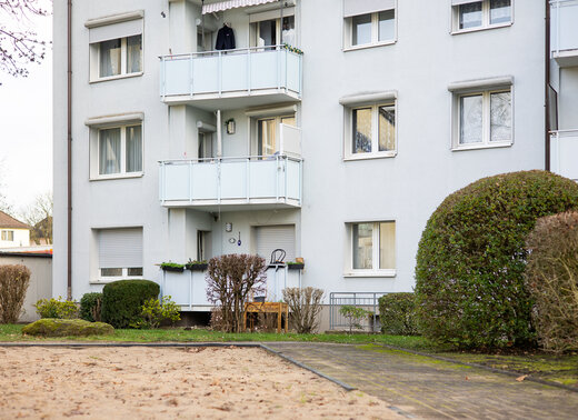 Mehrgeschossiges Mehrfamilienhaus mit heller Fassade, Balkonen und Fenstern, davor Grünflächen mit Sträuchern und einem Gehweg. Im Vordergrund ist die Sandfläche eines Spielplatzes zu sehen.