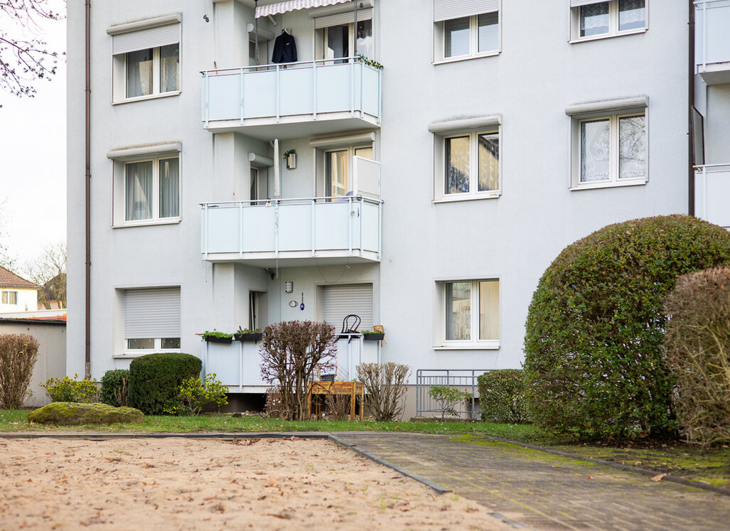 Mehrgeschossiges Mehrfamilienhaus mit heller Fassade, Balkonen und Fenstern, davor Grünflächen mit Sträuchern und einem Gehweg. Im Vordergrund ist die Sandfläche eines Spielplatzes zu sehen.