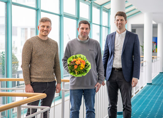 Drei Männer stehen in einem hellen Flur, einer von ihnen hält einen Blumenstrauß.