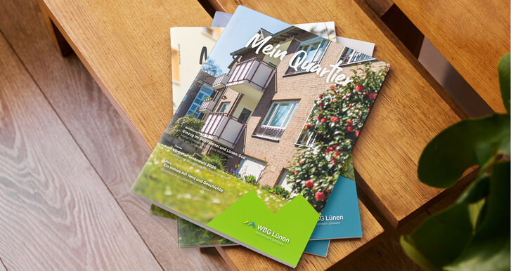 Das Mitgliedermagazin „Mein Quartier“ liegt gestapelt auf einer Holzbank. Auf dem Cover ist eine Wohnanlage mit Pflanzen im Vordergrund zu sehen, unten rechts befindet sich das WBG-Lünen-Logo mit dem Schriftzug „Gemeinsam zuhause“.