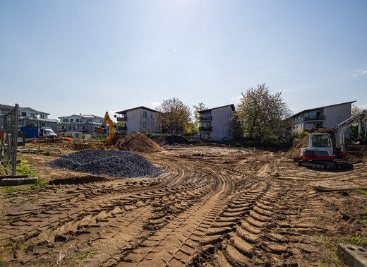 Baustelle mit frischer Baugrube, Erdhaufen, Schotter und zwei Baggern. Im Hintergrund sind Wohnhäuser zu sehen.