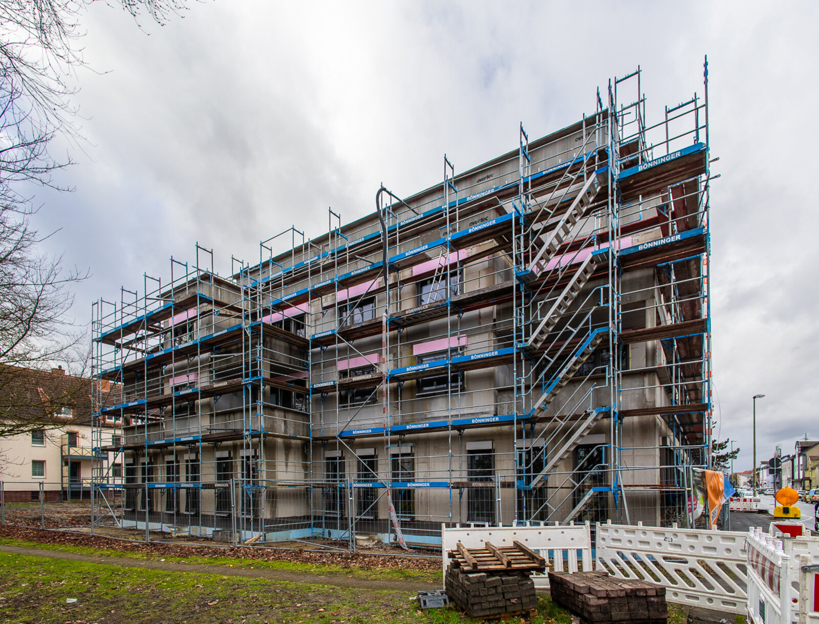 Neubau des Wohn- und Geschäftshauses in Lünen, vollständig von Baugerüst umgeben, mit Außentreppen am Gerüst und Baustellenabsperrungen im Vordergrund.