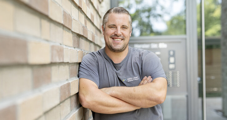 Porträt eines freundlichen Handwerkers der WBG Lünen, der mit verschränkten Armen vor einer Hauswand steht und in die Kamera lächelt. Im Hintergrund ist ein Hauseingang zu sehen.