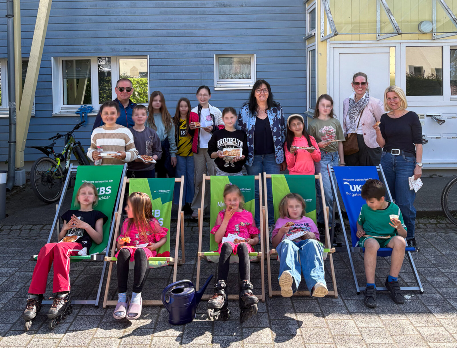 Gruppe aus Kindern und Erwachsenen steht und sitzt vor einem Wohnhaus, einige essen Pizza und lächeln in die Kamera.