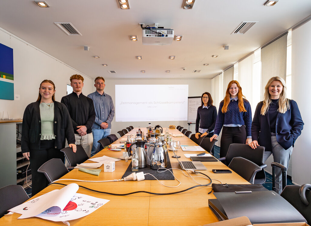 Auszubildende der WBG Lünen stehen gemeinsam mit der Seminarleitung in einem Seminarraum. Auf dem Konferenztisch liegen Seminarunterlagen, im Hintergrund ist eine Präsentation zu sehen.