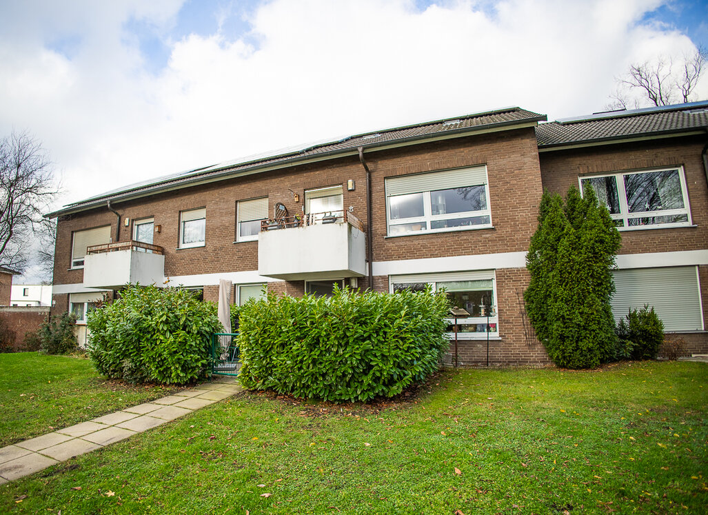 Auf dem Bild ist ein zweigeschossiges Mehrfamilienhaus mit brauner Klinkerfassade zu sehen. Das Gebäude verfügt über mehrere Balkone mit weißen Brüstungen sowie große Fenster mit Rollläden. Im Vordergrund erstreckt sich eine gepflegte Grünfläche mit Rasen und Hecken.