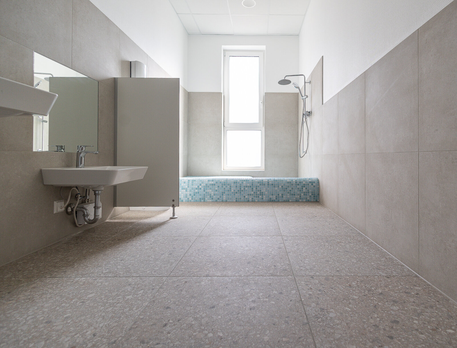 Badezimmer in der fertiggestellten Kindertagesstätte mit Dusche, Waschbecken und hellen Fliesen.