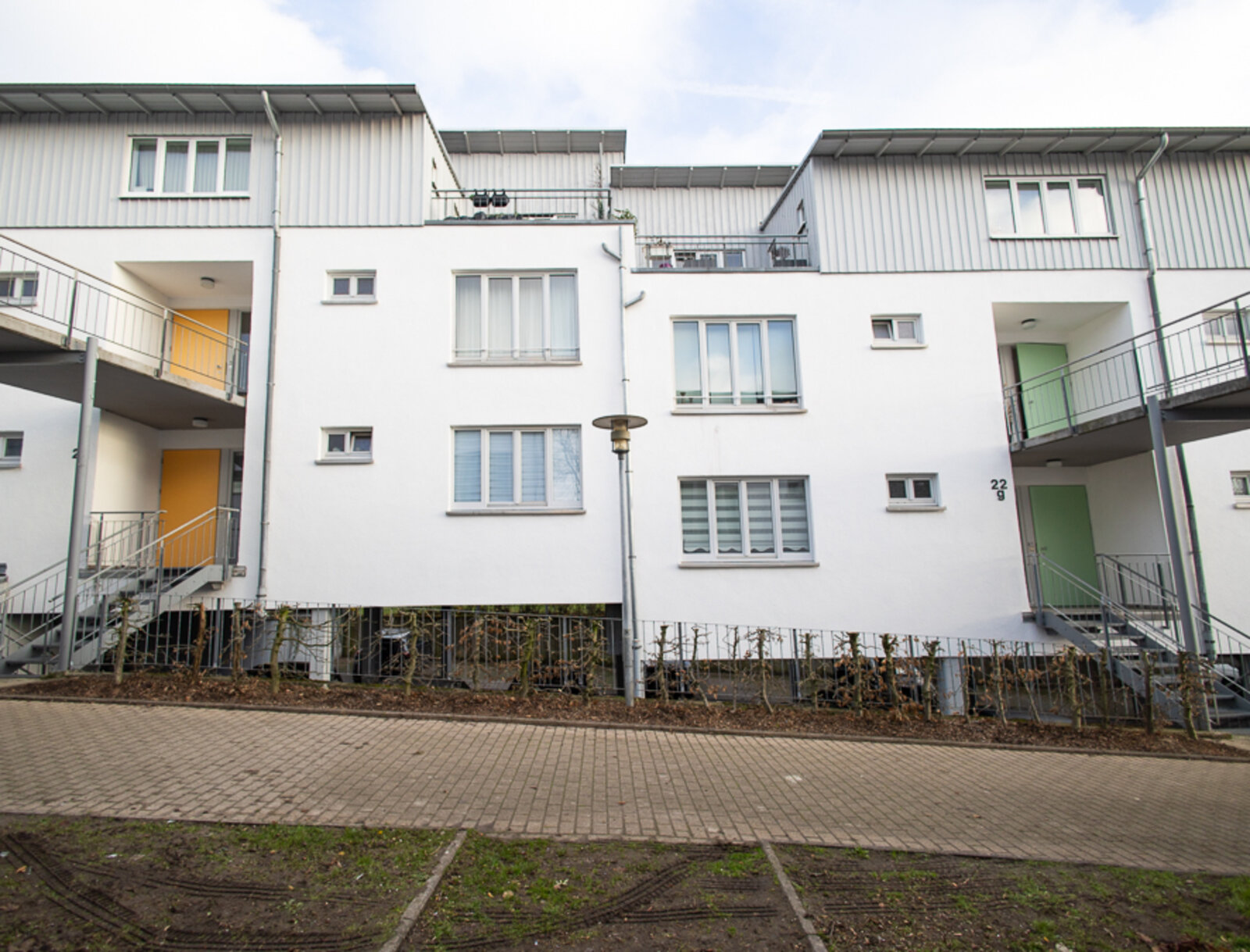 Frontansicht der Wohnanlage mit weißer Fassade, mehreren Fenstern und Außentreppen. Vor dem Gebäude verlaufen ein gepflasterter Weg und begrünte Flächen.