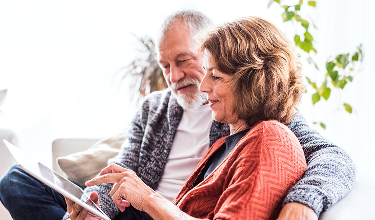 Halfpoint | Adobe Stock Ein Pärchen mittleren Alters sitzen auf dem Sofa und schauen gemeinsam auf ein Tablet.