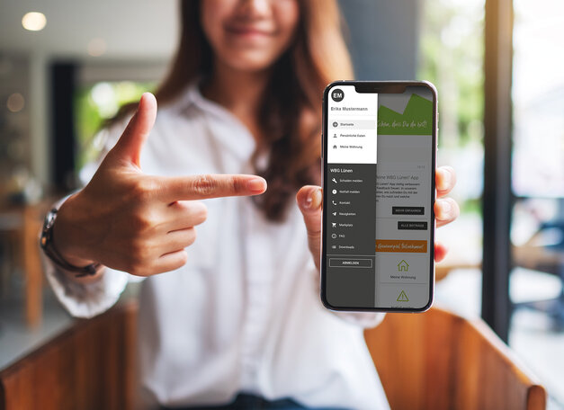 Weibliche Person zeigt auf ein Smartphone. Auf dem Display ist das Menü der App zu sehen.