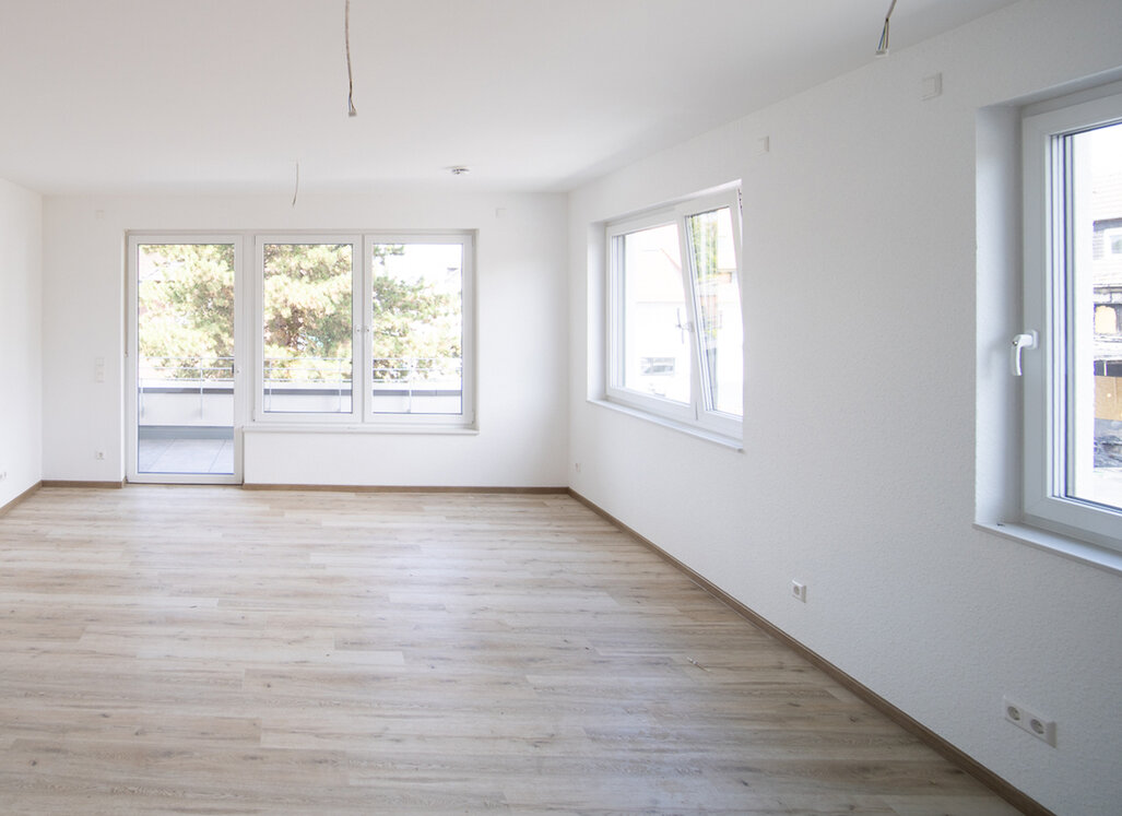 Blick in ein helles Wohnzimmer mit mehreren Fenstern und gegenüberliegendem Ausgang zum Balkon.