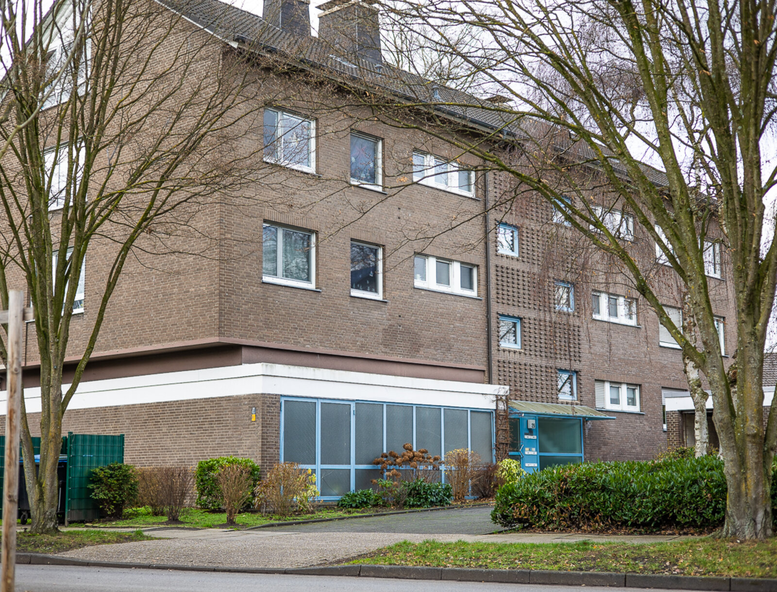 Mehrfamilienhaus mit Klinkerfassade, mehreren Fensterreihen und Eingangsbereich, umgeben von Bäumen und Grünflächen.