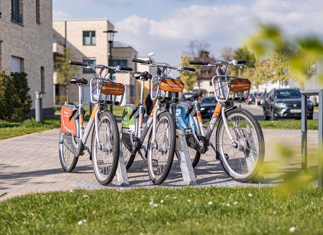 Mehrere Leihfahrräder stehen an einer Fahrradstation in einem Wohnquartier. Im Hintergrund sind Wohngebäude, parkende Autos und Grünflächen zu sehen.