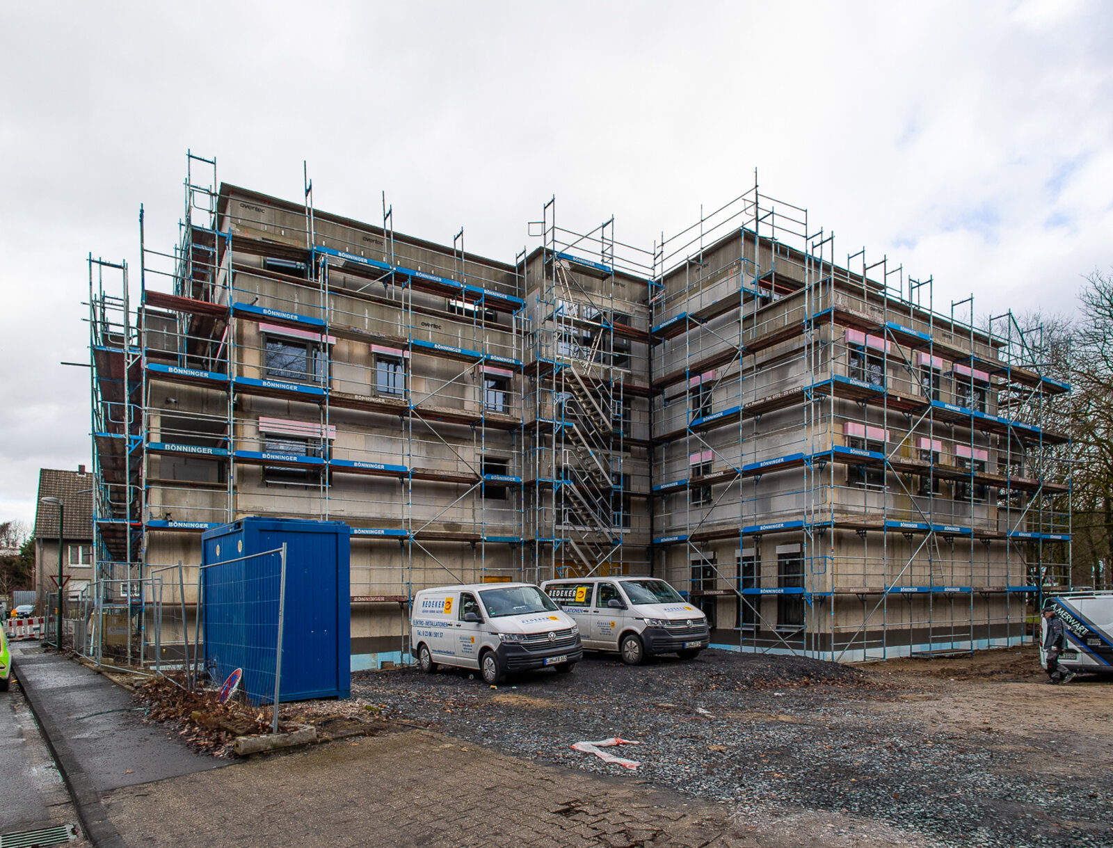 Neubau eines Mehrfamilienhauses an der Kleinen Bebelstraße in Lünen mit Baugerüst, Baustellencontainer und mehreren Handwerkerfahrzeugen auf dem Baugrundstück.