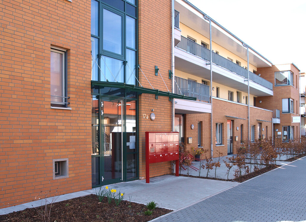 Mehrgeschossiges Wohngebäude mit brauner Klinkerfassade, Balkonen und Eingangsbereich mit Briefkastenanlage entlang eines gepflasterten Weges.