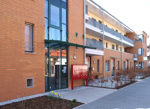 Mehrgeschossiges Wohngebäude mit brauner Klinkerfassade, Balkonen und Eingangsbereich mit Briefkastenanlage entlang eines gepflasterten Weges.