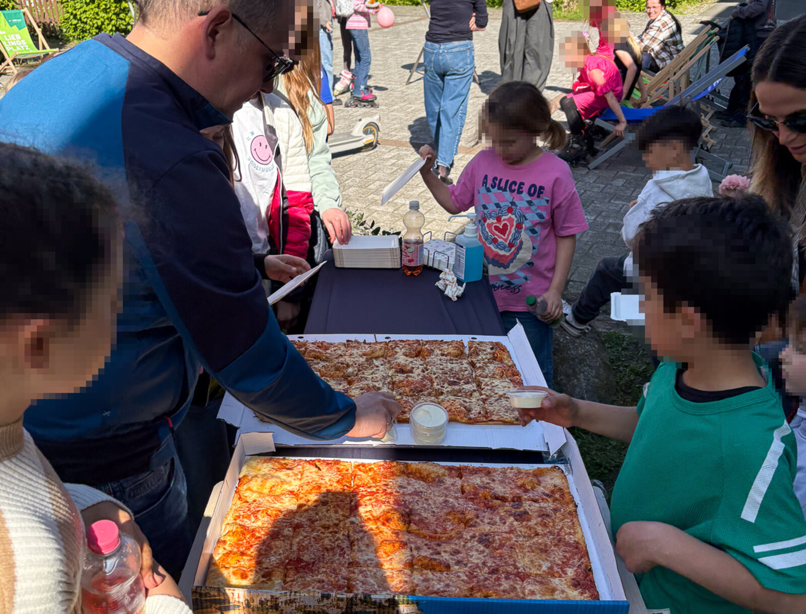 Kinder und Erwachsene stehen im Freien um einen Tisch und nehmen sich Pizza aus großen Kartons.