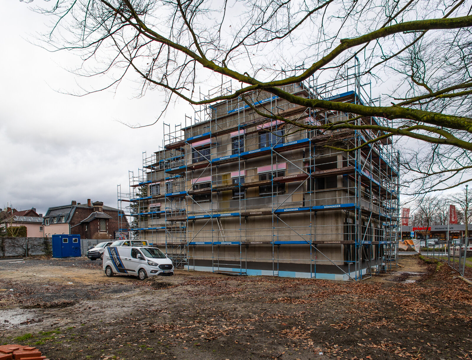 Vollständig eingerüstetes Mehrfamilienhaus, parkendem Handwerkerfahrzeug auf dem unbefestigten Grundstück und kahlen Baumästen im Vordergrund.
