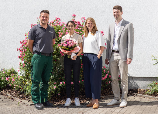 Vier Mitarbeitende der WBG Lünen stehen vor blühenden Rosen. Eine junge Frau wird mit einem Blumenstrauß zur erfolgreich bestandenen Ausbildung beglückwünscht.