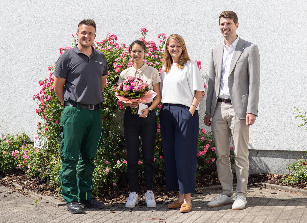 Vier Mitarbeitende der WBG Lünen stehen vor blühenden Rosen. Eine junge Frau wird mit einem Blumenstrauß zur erfolgreich bestandenen Ausbildung beglückwünscht.