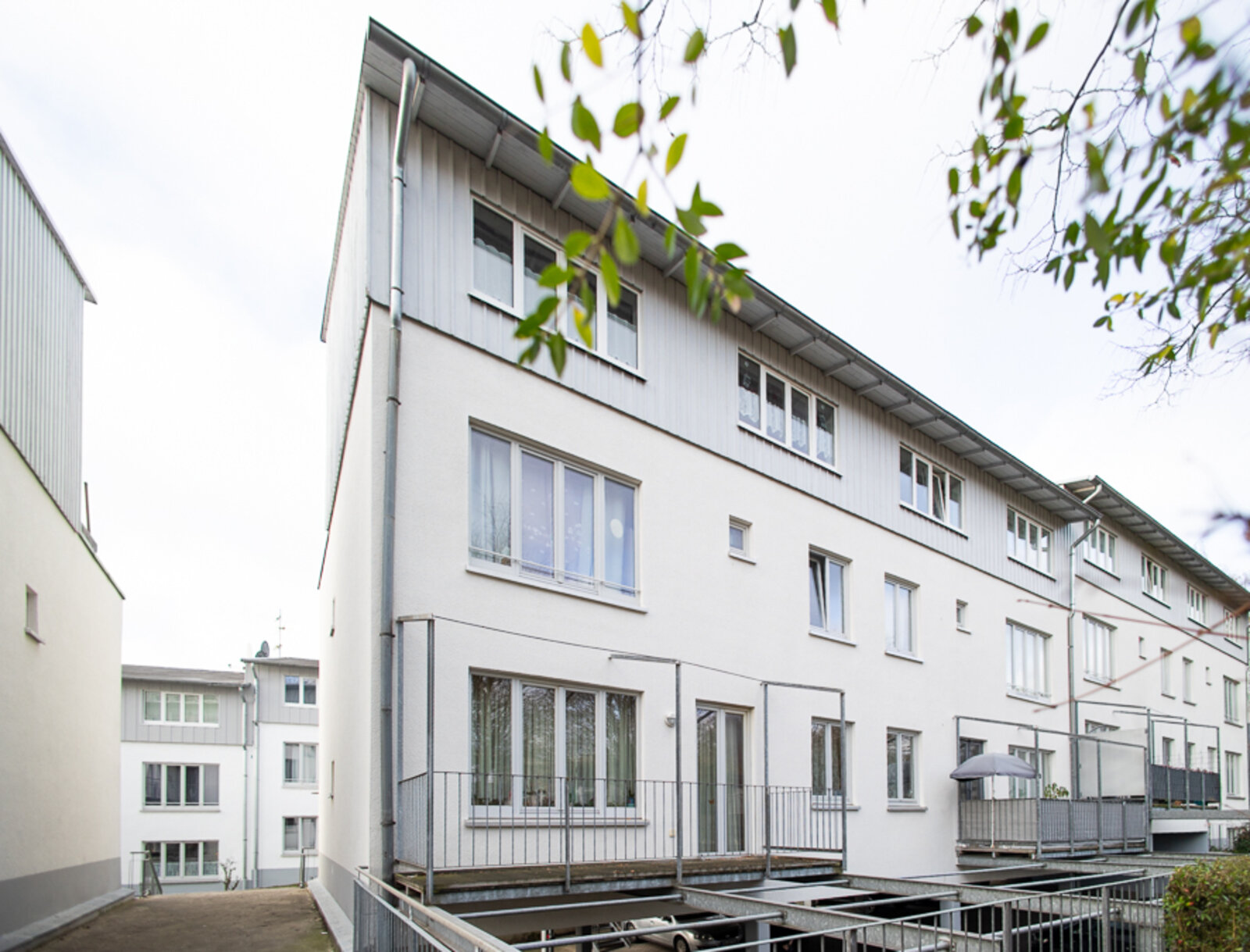 Rückansicht der Wohnanlage mit heller Fassade, großen Fenstern und Balkonen. Es sind Geländer und Stellplätze unter dem Gebäude zu sehen.