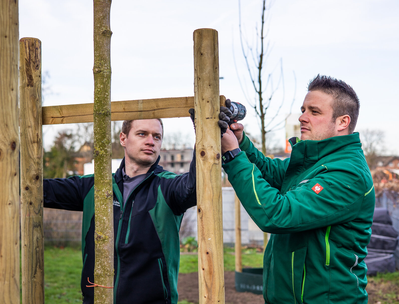 Zwei Mitarbeitende der WBG Lünen in grüner Arbeitskleidung befestigen mit einem Akkuschrauber eine Holzstütze an einem frisch gepflanzten Baum. Im Hintergrund ist ein weiterer junger Baum und eine Grünfläche zu sehen.