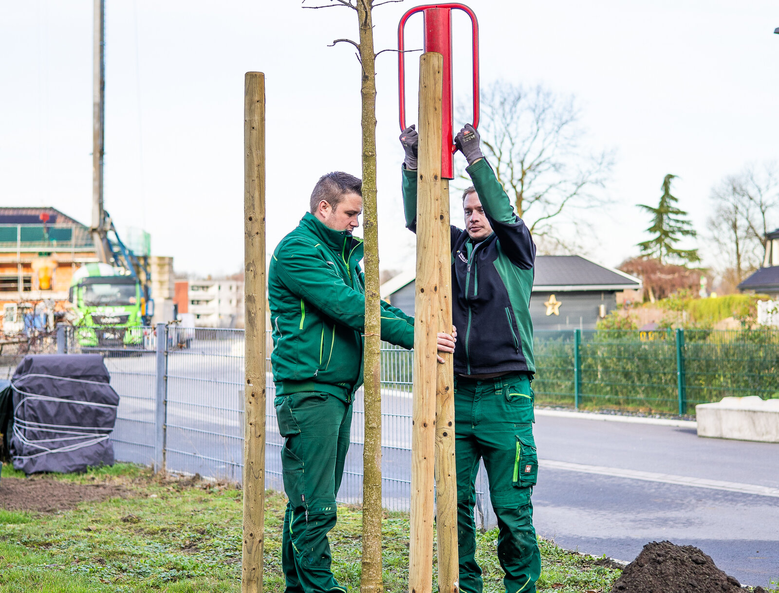 Zwei Mitarbeitende der WBG Lünen pflanzen einen jungen Baum auf einer Grünfläche und positionieren die Holzstützen in der Erde. Im Hintergrund sind eine Straße, Baugeräte und Wohngebäude zu sehen.