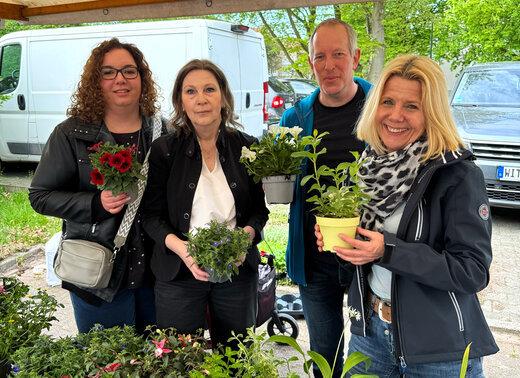 Mitarbeitende der VIVAWEST und der WBG Lünen stehen gemeinsam an einem Pflanzstand und halten verschiedene Blumen und Pflanzen in Töpfen. Im Vordergrund sind weitere Pflanzen zu sehen.