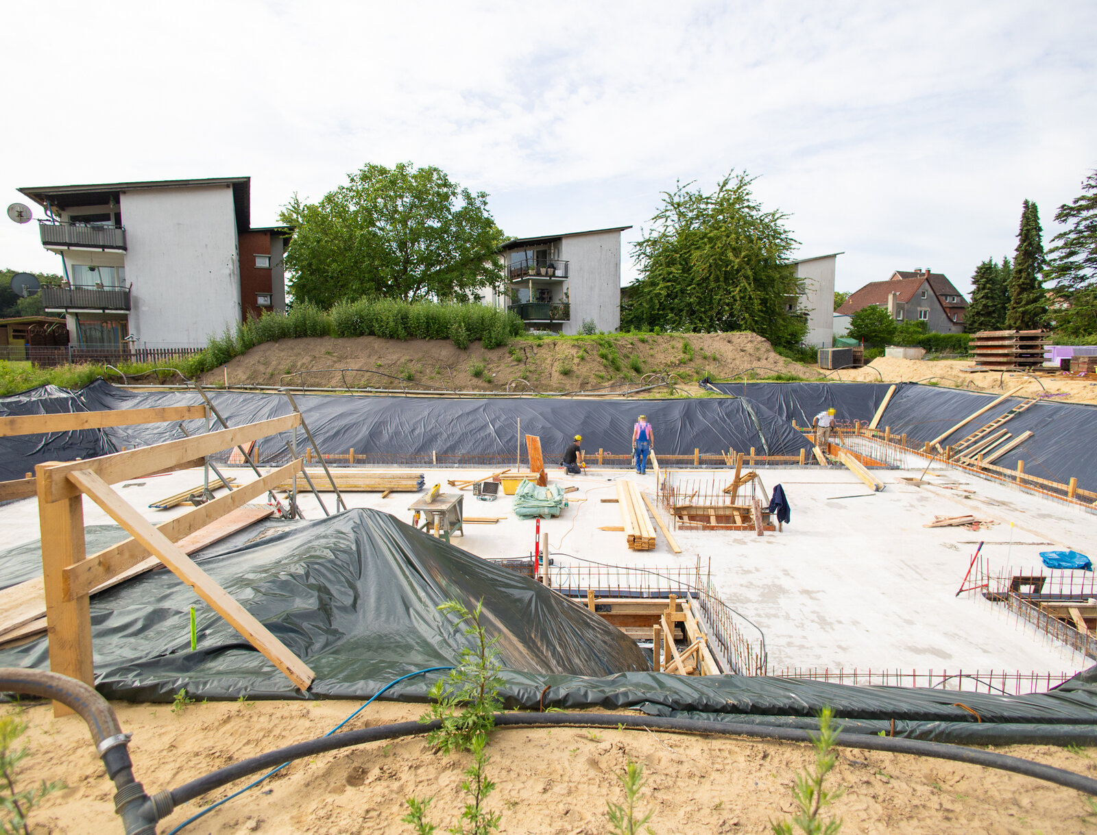 Baugrube des Neubauprojekts mit fertiggestellter Bodenplatte. Auf der Baustelle sind Baumaterialien, Schalungen und mehrere Handwerker*innen bei den weiteren Vorbereitungen zu sehen.