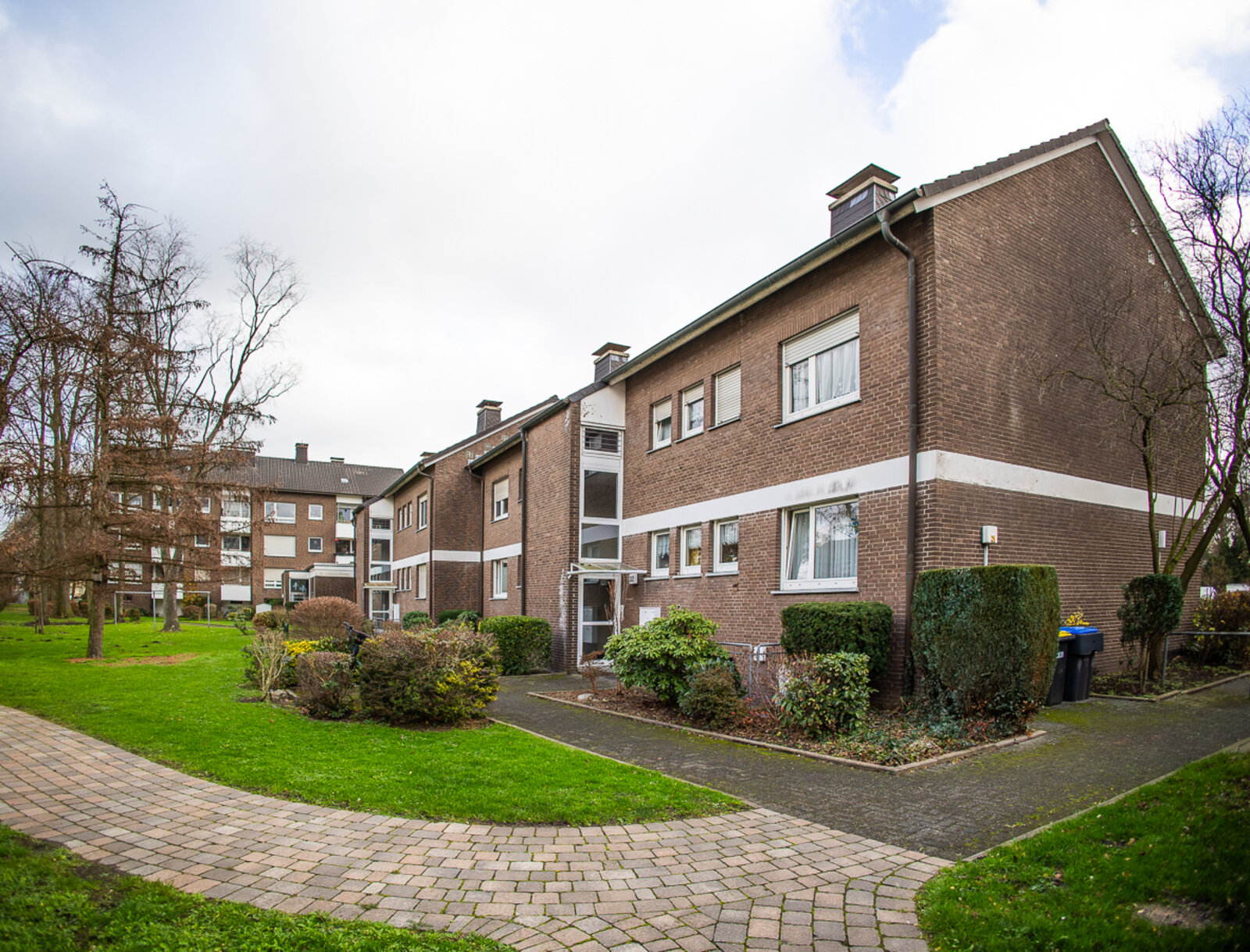 Das Bild zeigt die Rückansicht eines zweigeschossigen Mehrfamilienhaus mit Klinkerfassade, Balkonen und großen Fenstern. Davor liegt eine gepflegte Grünfläche mit Rasen, Hecken und Gehwegen, die den ruhigen Wohnhof umrahmen.