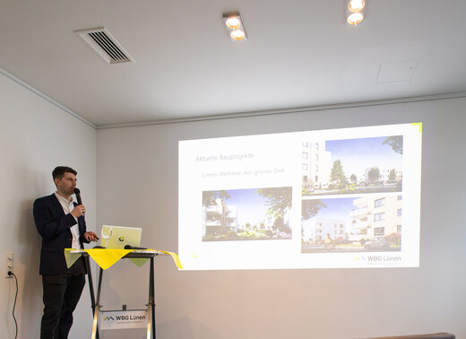 Vorstand Jan Hische steht an einem Stehtisch und spricht zu den Mitarbeitenden der WBG Lünen. Im Hintergrund läuft eine PowerPoint-Präsentation.
