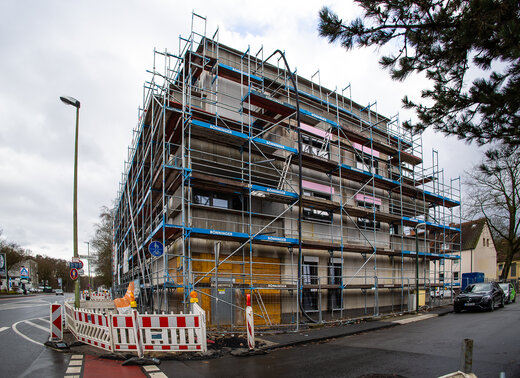 Mehrgeschossiges Wohngebäude, vollständig eingerüstet, mit Baustellenabsperrungen und Straßenkreuzung im Vordergrund.