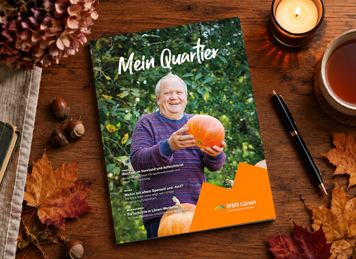 Das Mitgliedermagazin „Mein Quartier“ liegt auf einem herbstlich geschmücktem Tisch. Auf dem Cover ist ein Mann zu senden, der einen Kürbis in den Händen hält und in die Kamera lächelt. Unten rechts ist das WBG-Lünen-Logo mit dem Schriftzug „Gemeinsam zuhause“ abgebildet.