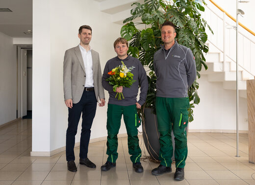 Neuer Mitarbeiter Sven Riedel mit Blumenstrauß gemeinsam mit Vorstand Jan Hische und Hendrik Kleinbecker im Eingangsbereich der Verwaltung.