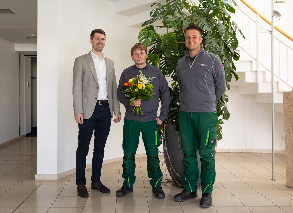 Neuer Mitarbeiter Sven Riedel mit Blumenstrauß gemeinsam mit Vorstand Jan Hische und Hendrik Kleinbecker im Eingangsbereich der Verwaltung.