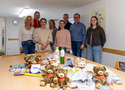 Gruppenfoto von acht Personen in den Räumlichkeiten der Hausaufgabenhilfe. Auf dem Tisch liegen Give Aways wie Stofftiere, Trinkflaschen und Pixie Bücher.