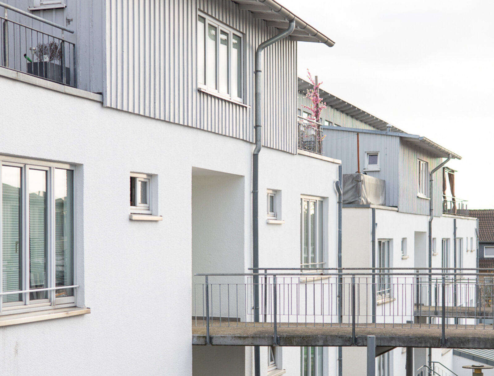 Mehrgeschossige Wohnhäuser mit hellen Fassaden und grauen Balkon- und Fassadenelementen. Zu sehen sind Balkone, Fenster und ein Laubengang mit Geländern